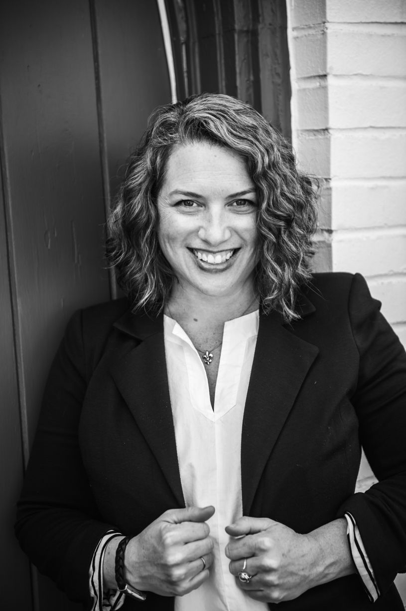 Professional headshot of a woman in a black blazer and white blouse against a brick wall.