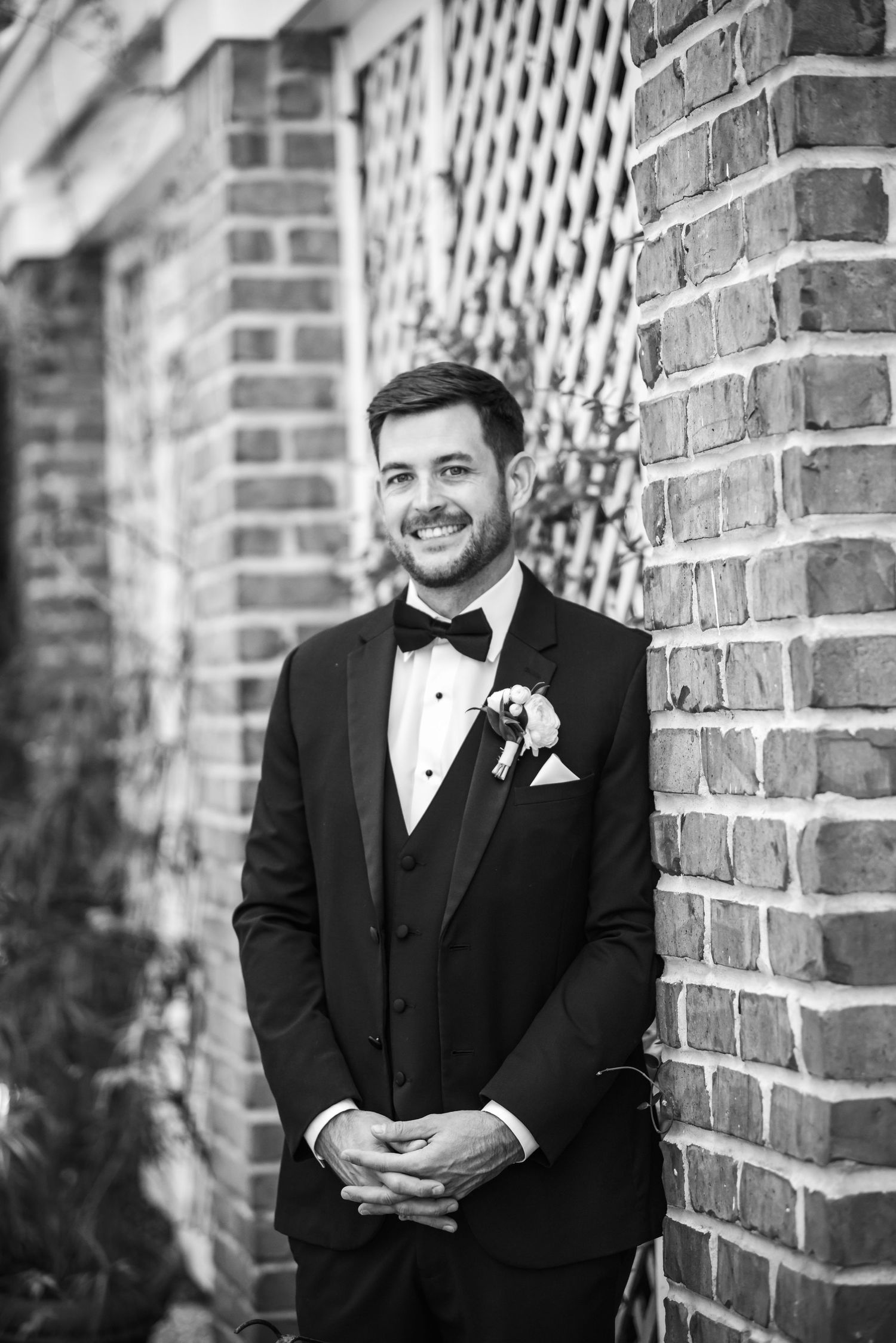 Smiling groom in black tuxedo with boutonniere leans against brick wall in elegant black and white wedding portrait.