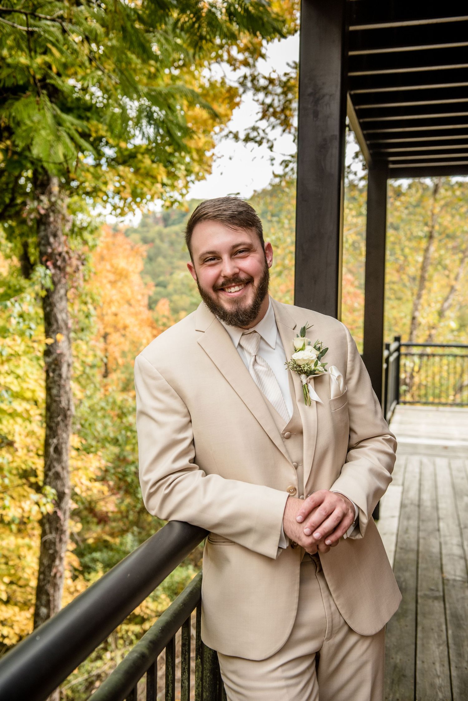 Smiling groom in beige suit with boutonniere poses on wooden deck surrounded by colorful autumn foliage.