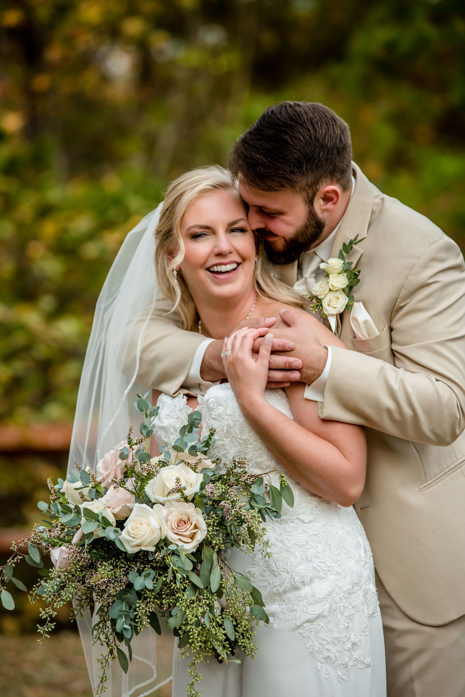 Bride and groom embrace outdoors, she holds white floral bouquet, he wears tan suit, autumn foliage background.