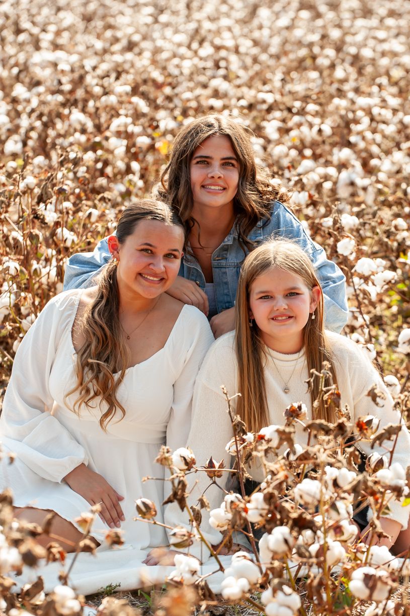 Three sisters pose together in a cotton field wearing coordinated white outfits.