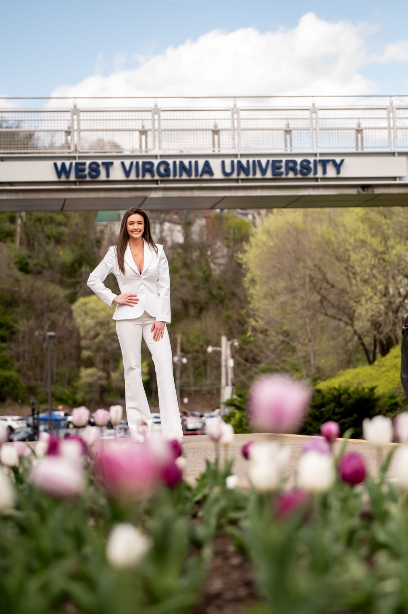 A scenic view of West Virginia University campus with blooming tulips in the foreground with a senior grad posing in front of it.