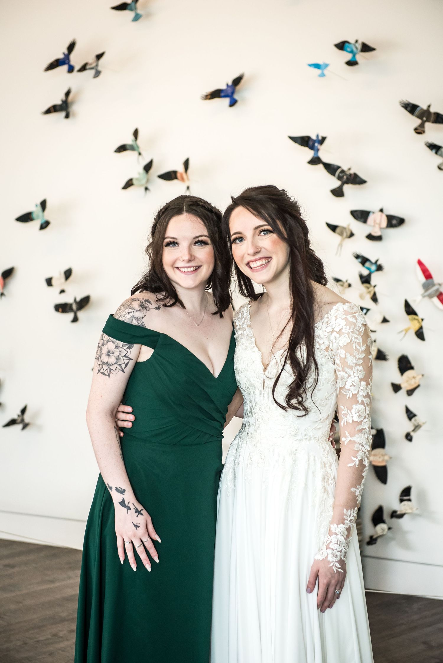 Two women pose together, one in a green gown and one in a white lace bridal dress, in front of a bird wall art.