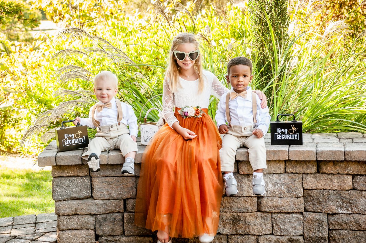 Three children sit on a stone wall outdoors, two boys in suspenders holding chalkboard signs and a girl in an orange tulle skirt.