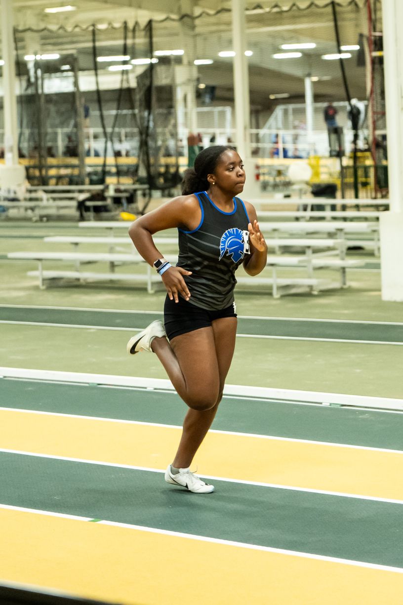 Track athlete in black uniform runs on indoor track with determined expression.