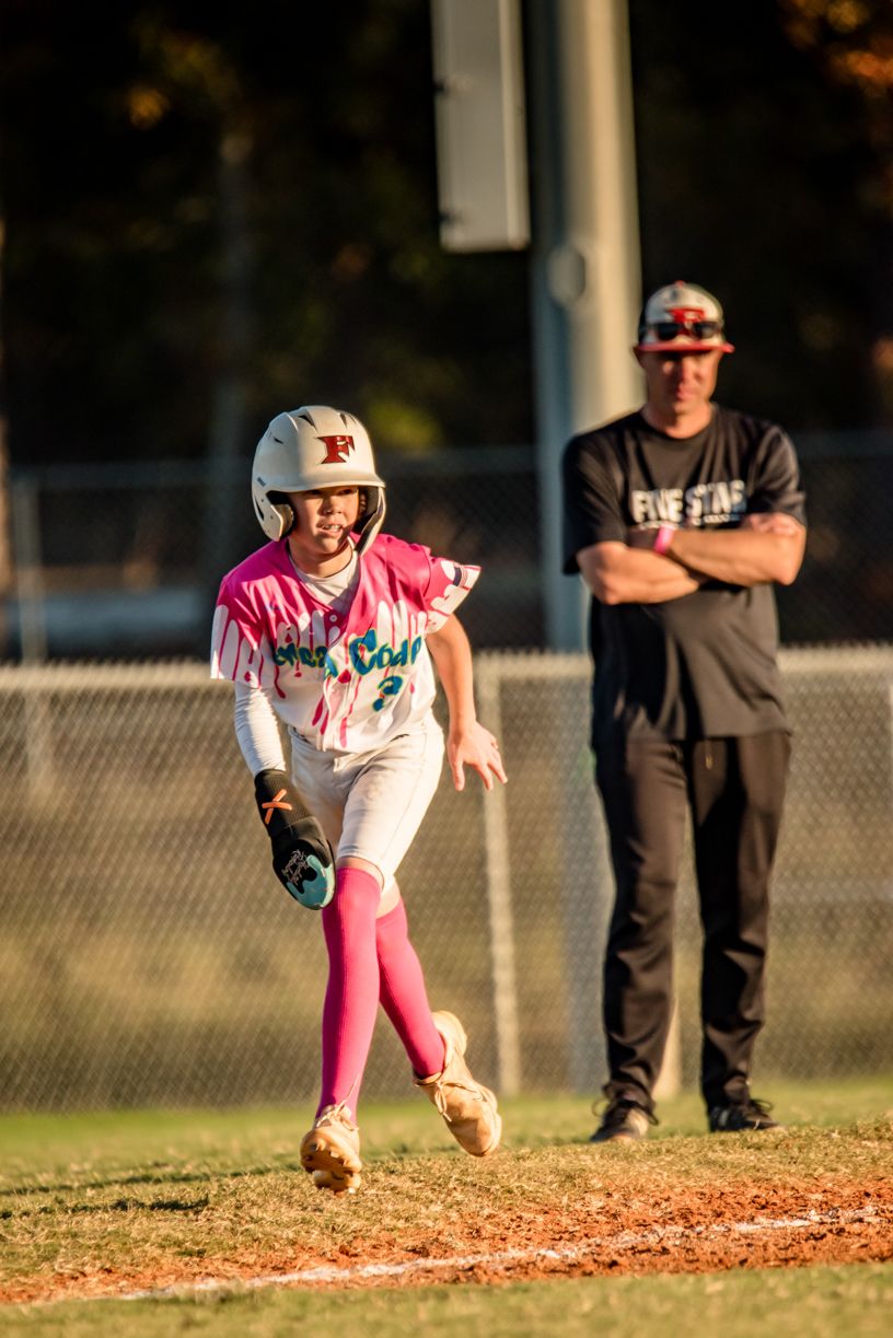Youth baseball player in pink uniform pitching on dirt mound during sunset game.
