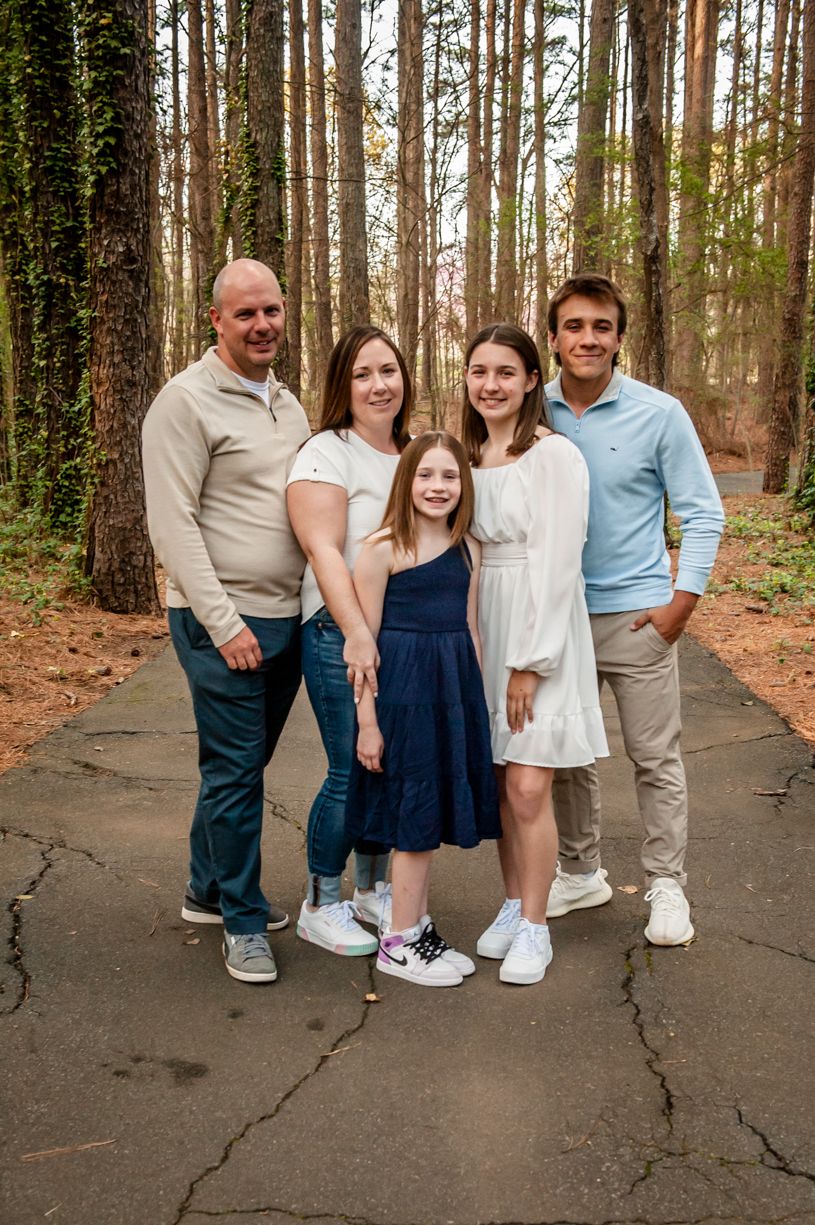 A family poses together on a wooded path wearing coordinated casual outfits in light colors.