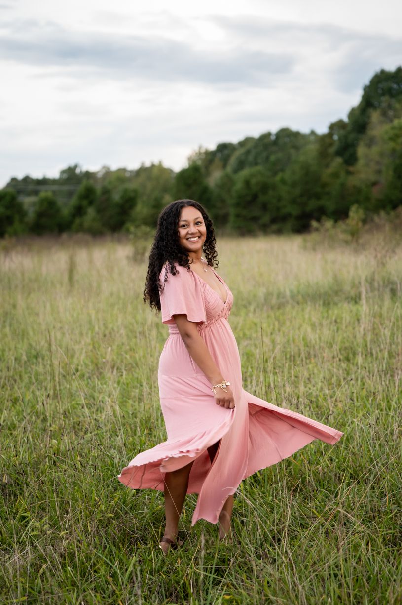 Senior girl in flowing pink dress twirls joyfully in a sunlit meadow of tall grass.