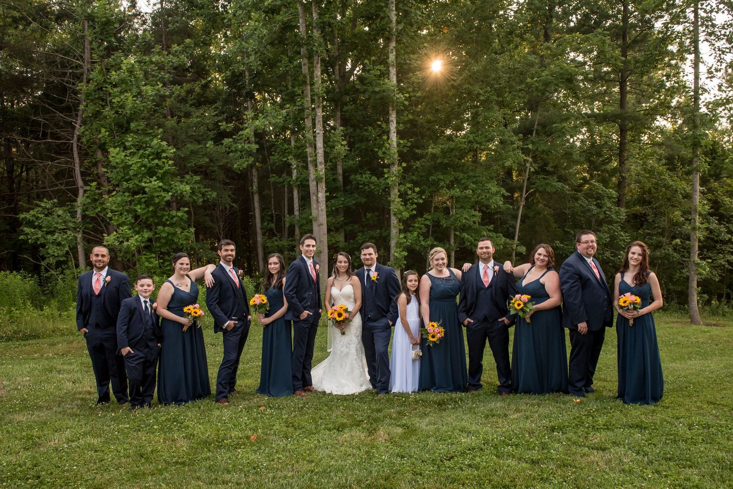 Wedding party in navy attire poses on green lawn with tall trees and golden sunset light in background.