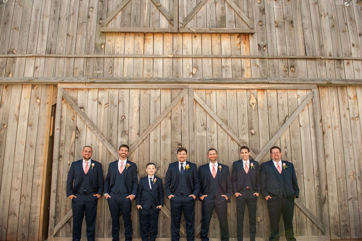 Groomsmen in navy suits with pink ties pose in front of rustic wooden barn doors at a wedding.
