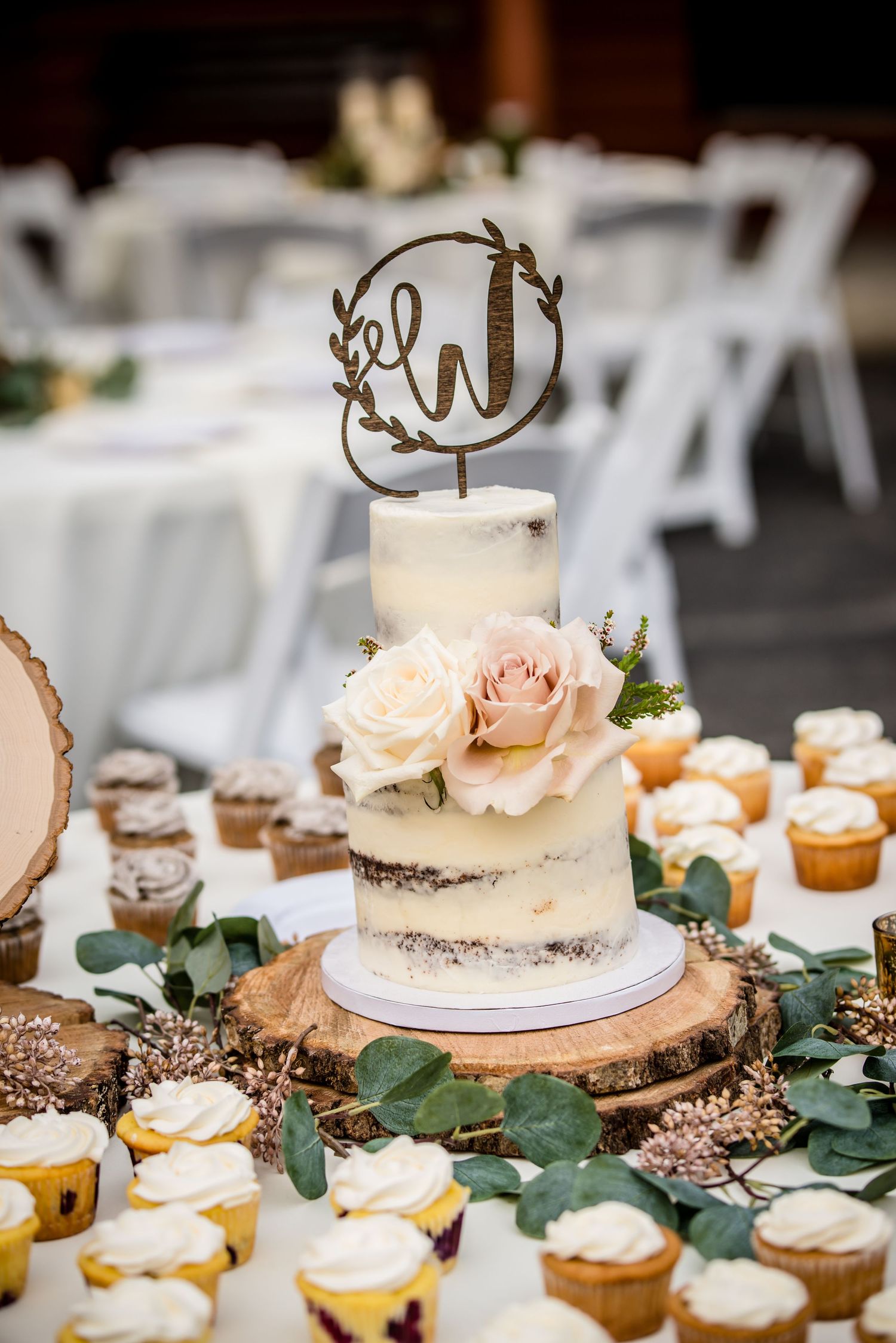 Two-tier semi-naked wedding cake with blush roses and monogram topper on wood slice, surrounded by cupcakes and greenery.