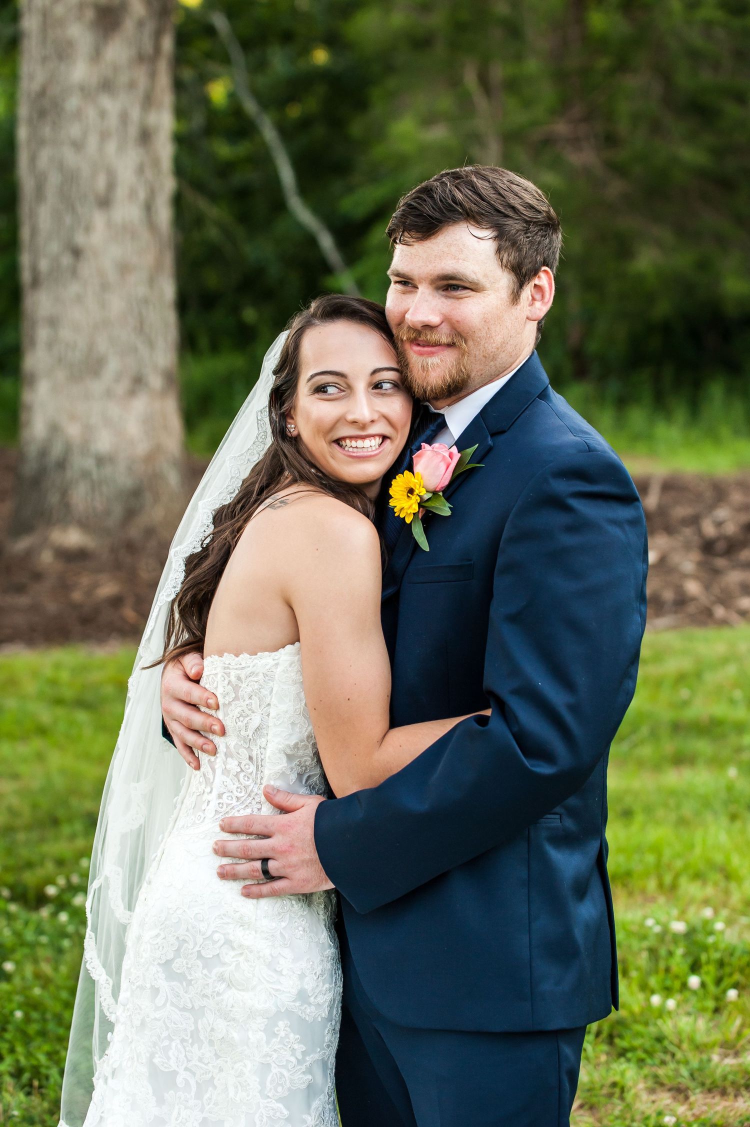 Bride in lace gown and veil embraces groom in navy suit with floral boutonniere outdoors at wedding.