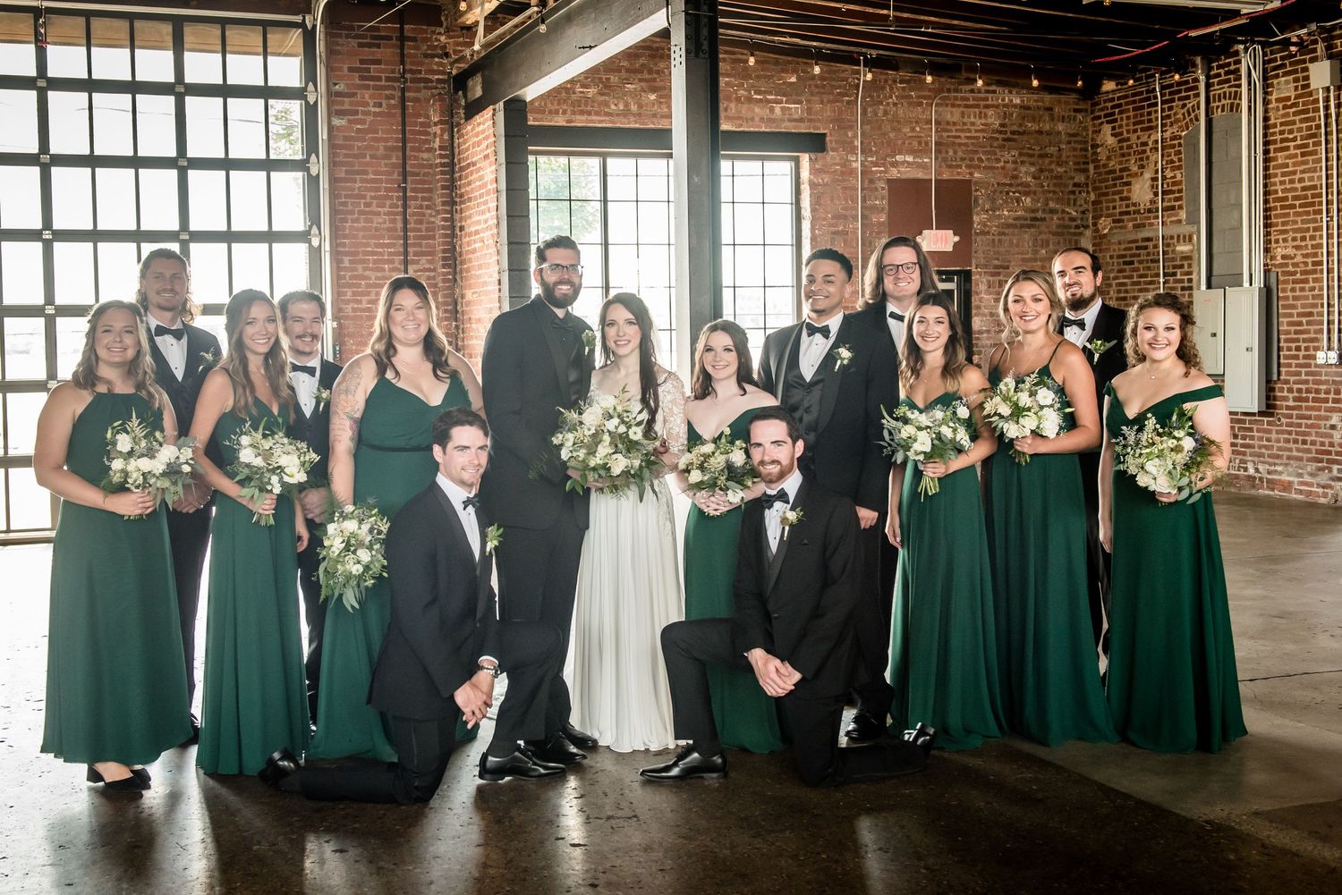 Wedding party in emerald green gowns and black tuxedos posing together in a rustic brick venue.