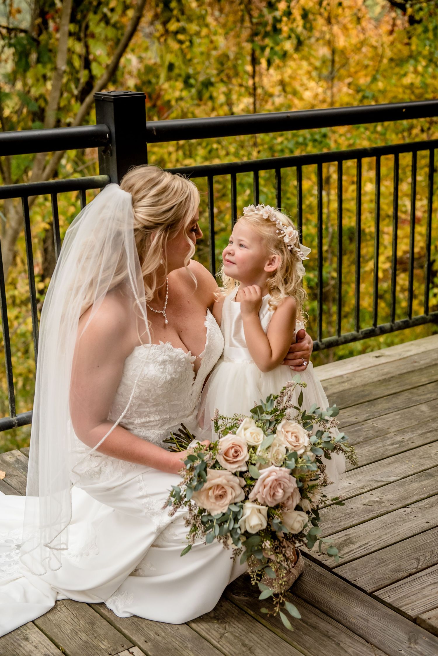 Bride in white gown and veil sits with flower girl in white dress on wooden deck, holding blush rose bouquet.