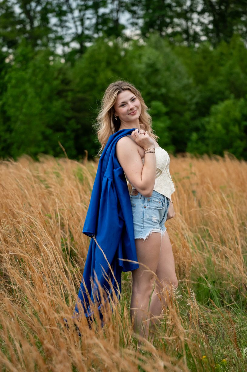 Summer portrait session of senior girl in tall golden grass with a flowing blue graduation cap over her shoulder.