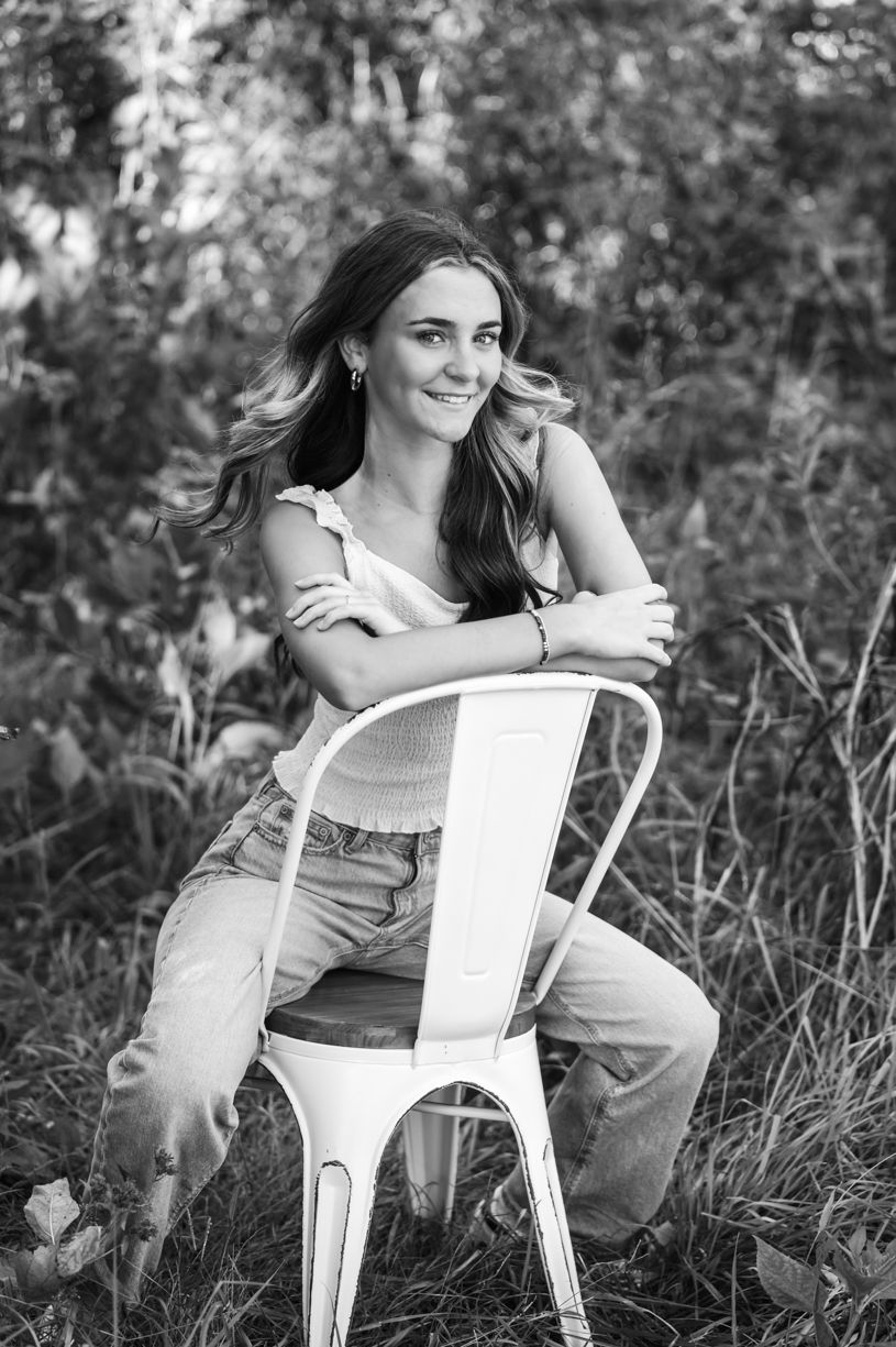Black and white portrait of a senior girl sitting on  a white metal chair in tall grass.