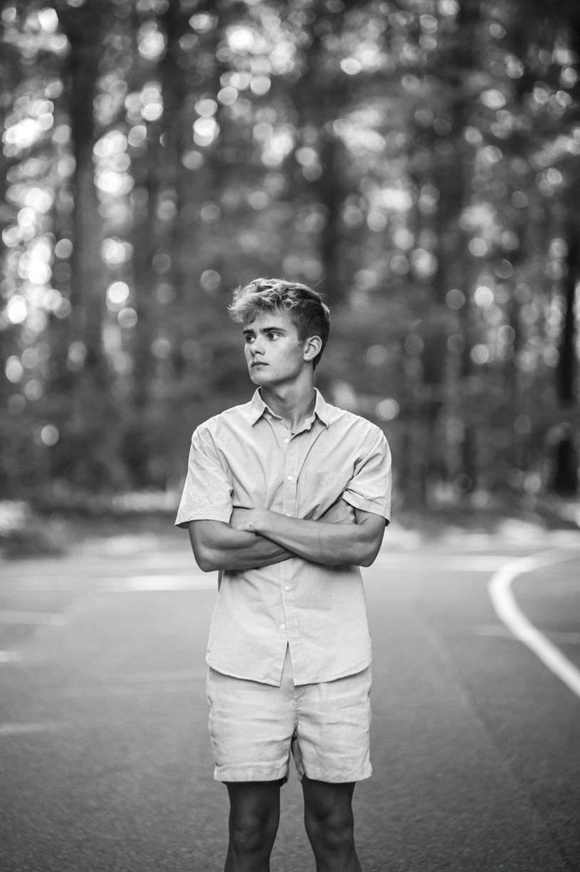 A senior guy folding his arms poses thoughtfully on forest road in black and white.