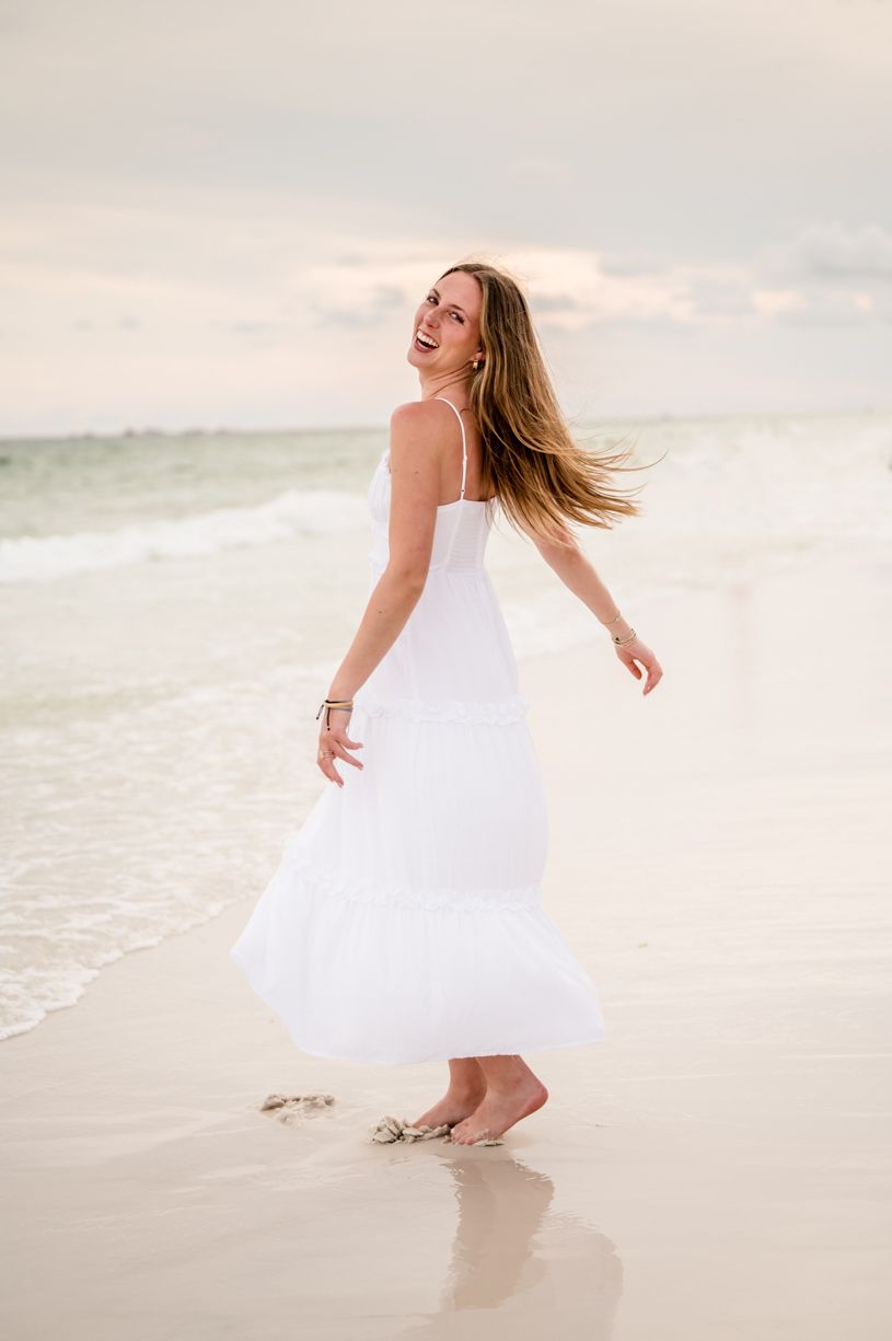 A joyful scene of a senior girl twirling in a white dress on a beach at sunset.