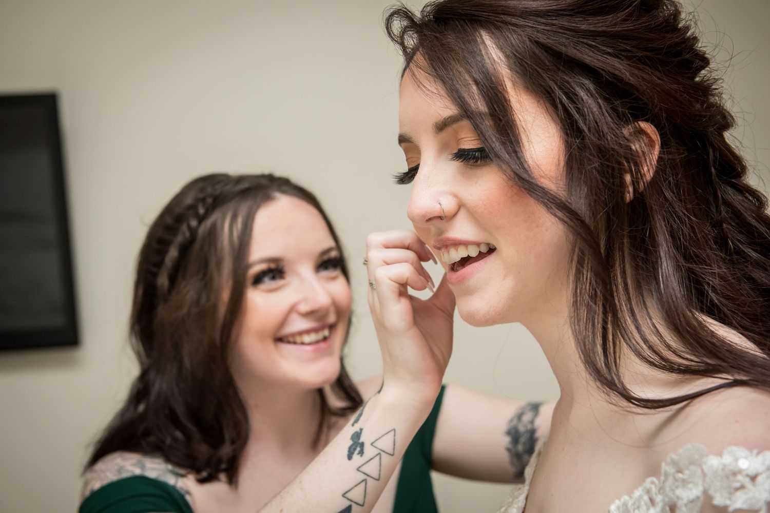 Bride smiling as bridesmaid in green dress helps with makeup during wedding preparations.