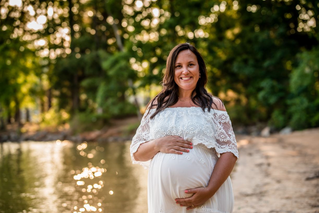 Maternity portrait of person in white lace dress standing by water at sunset.