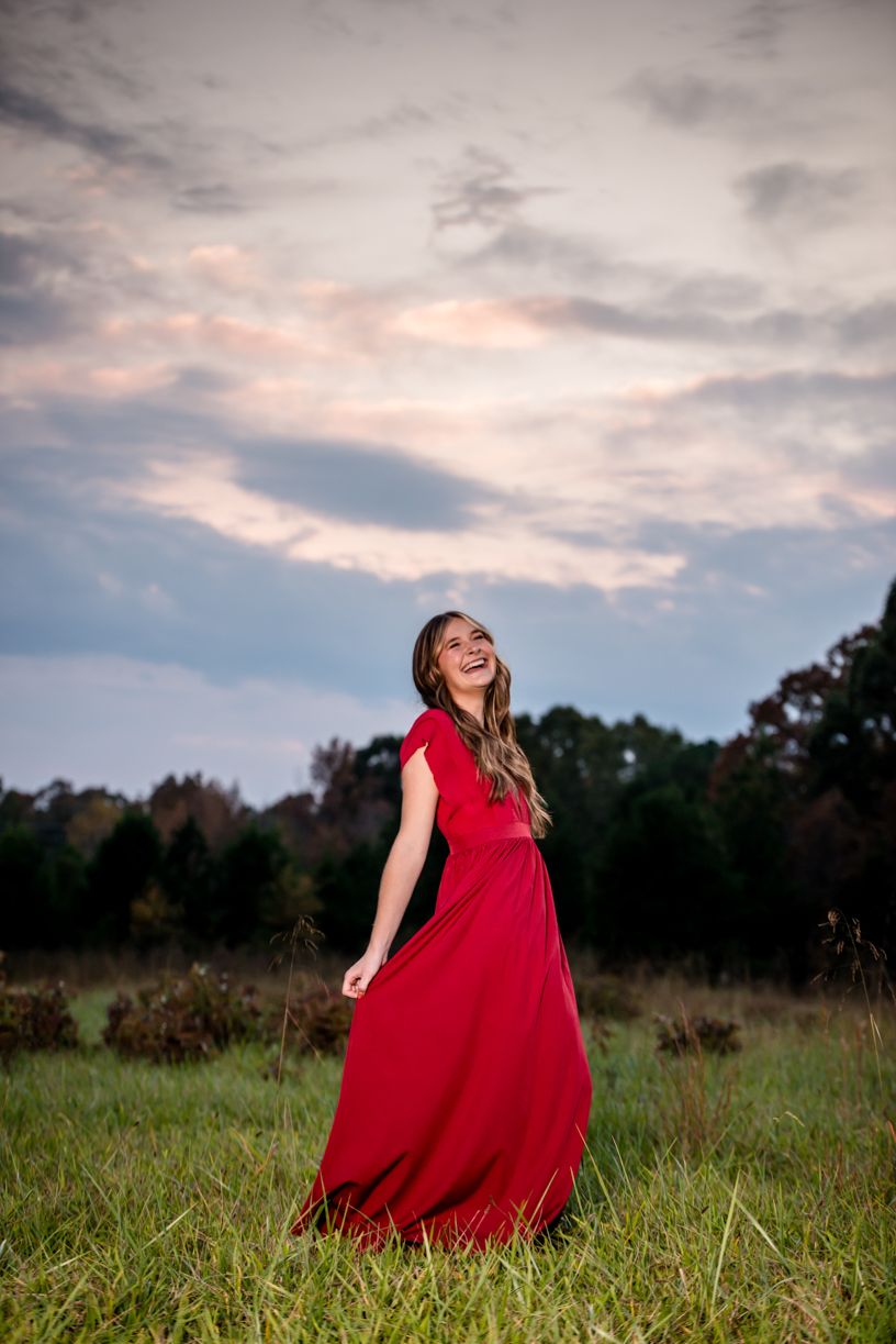 Senior girl in a dramatic red dress twirls in an open field against a moody sky at dusk.