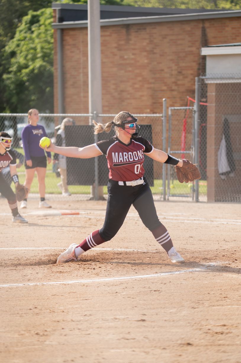 Softball pitcher in maroon uniform mid-delivery during game on dirt field.