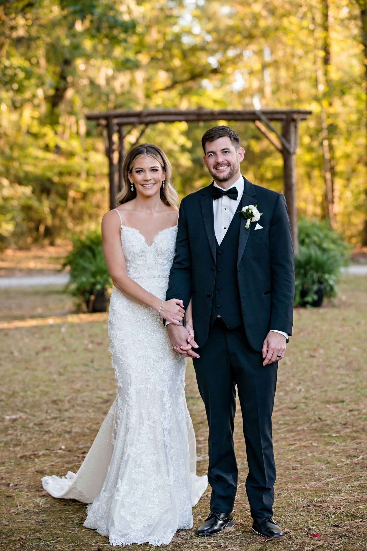 Bride in lace gown and groom in black tuxedo holding hands outdoors at a fall wedding ceremony.