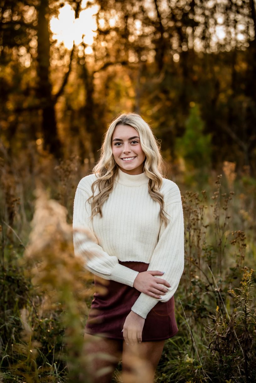 A senior girl standing in a field at sunset wearing a cream sweater.