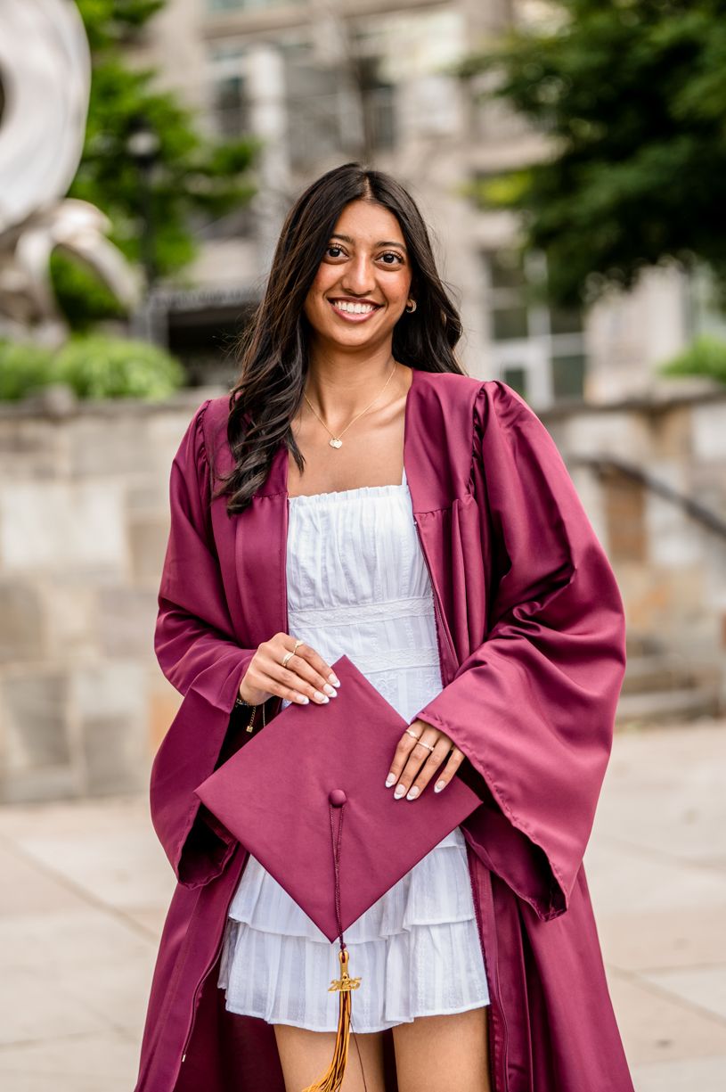 A graduate in a burgundy graduation gown posing on campus.