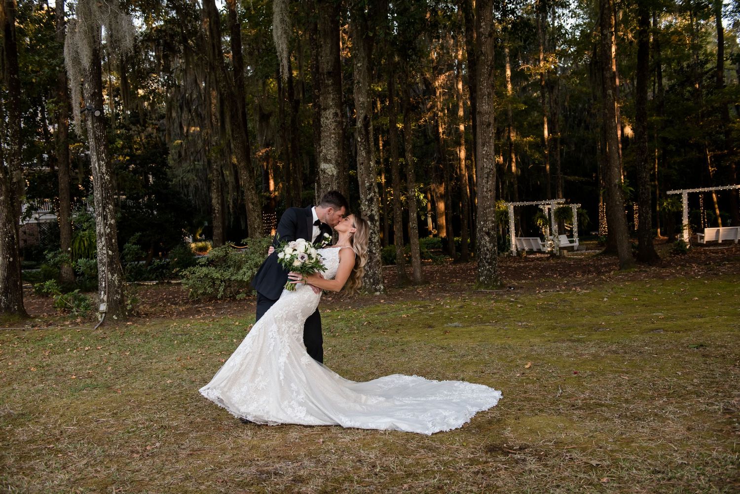 Bride and groom share a romantic dip kiss on a lush green lawn surrounded by tall trees at an outdoor wedding.