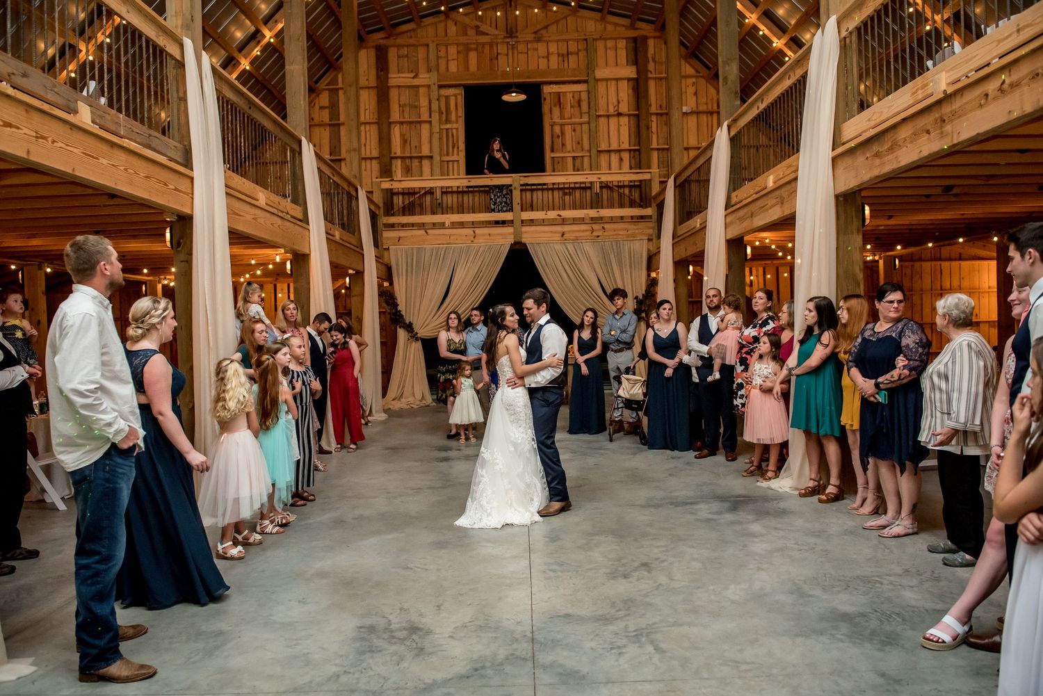 Bride and groom share first dance in rustic barn wedding venue surrounded by guests in formal attire.