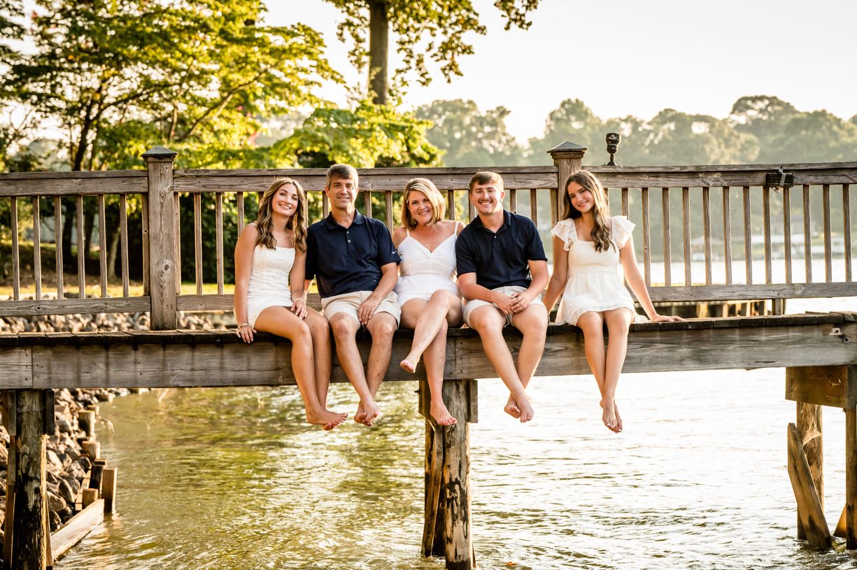 Family of 5 sitting casually on a wooden dock by the water at sunset.