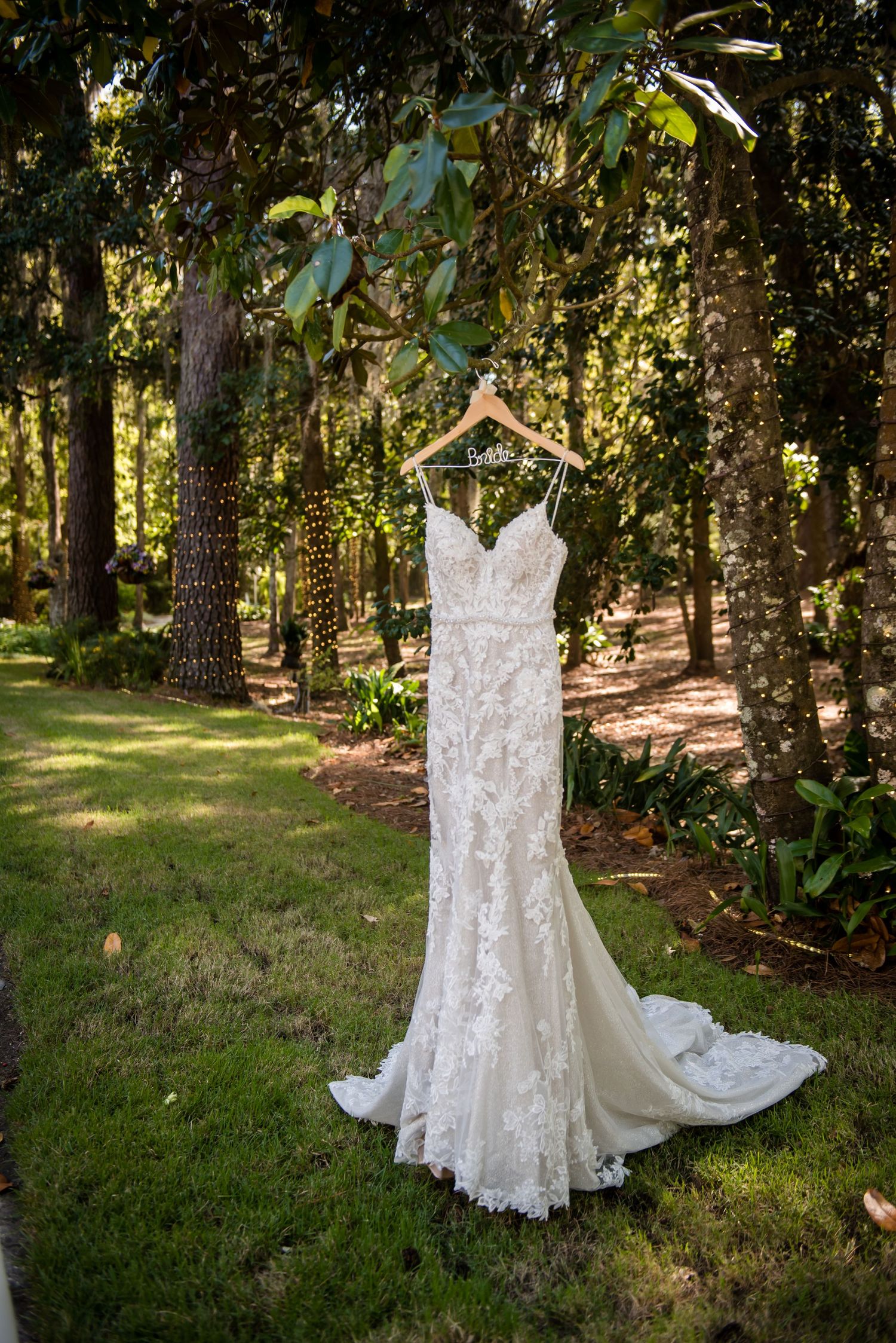 Elegant lace wedding dress with floral appliques hanging on wooden hanger outdoors in lush green forest setting.
