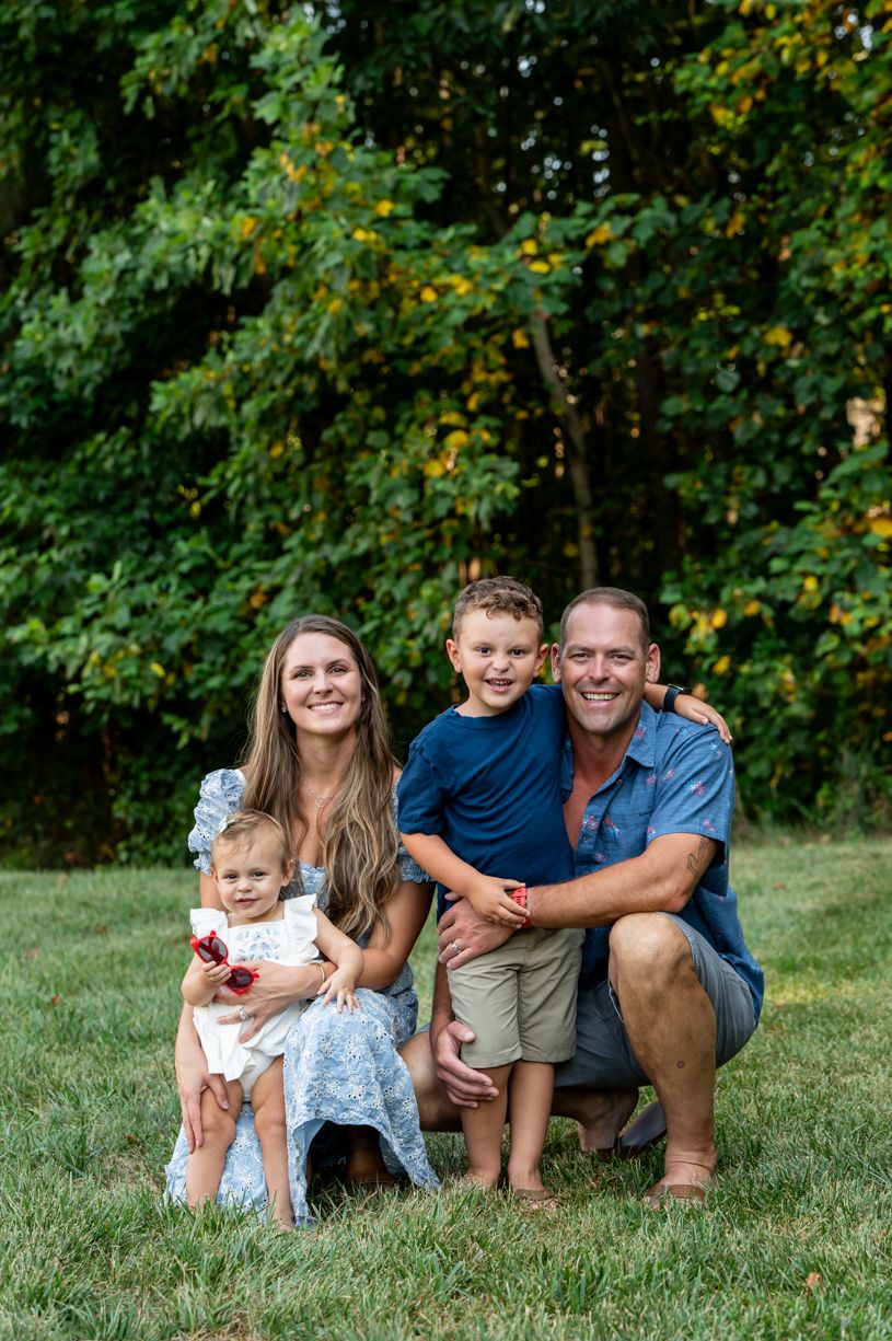 A family of 4 poses together on grass with trees in the background during summer.