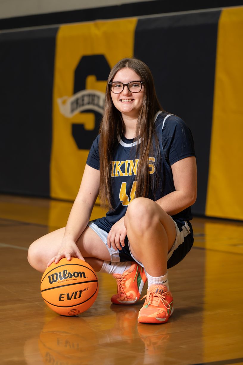 Basketball player in navy Vikings uniform kneeling with Wilson basketball on wooden court with yellow and black backdrop.