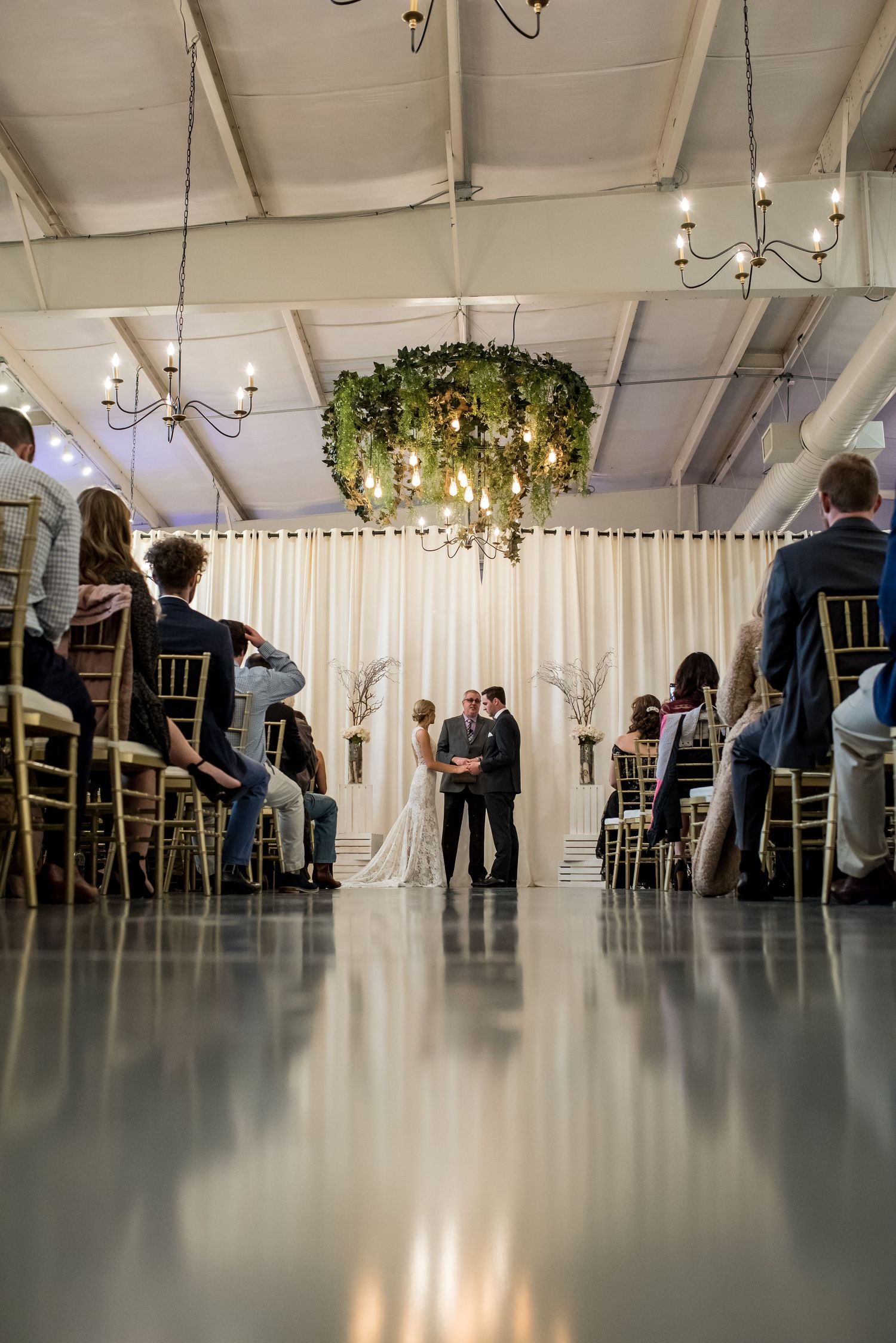 Indoor wedding ceremony with couple at altar, white drape backdrop, chandelier with greenery, guests seated in gold chairs.