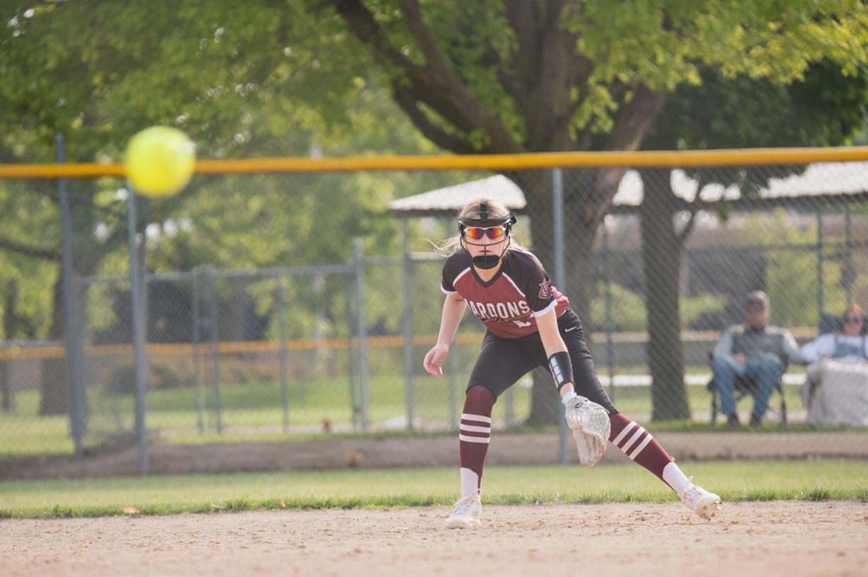 Softball player in maroon uniform fielding yellow ball on dirt infield.