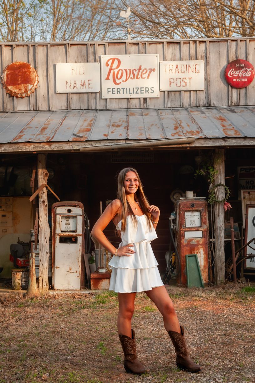 Rustic photoshoot in front of vintage store signs and weathered wooden buildings featuring a senior girl in a white dress.