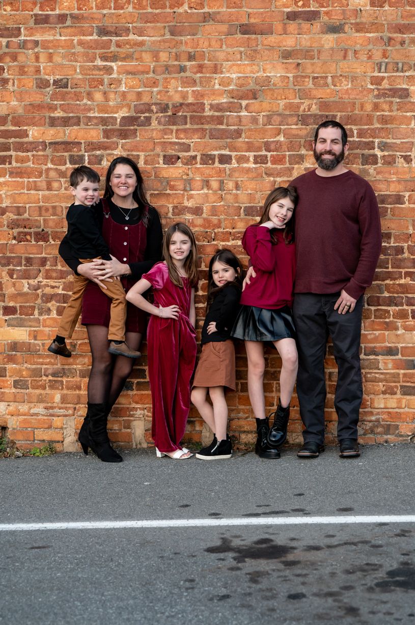 Family posing casually against a rustic brick wall wearing coordinated burgundy outfits.