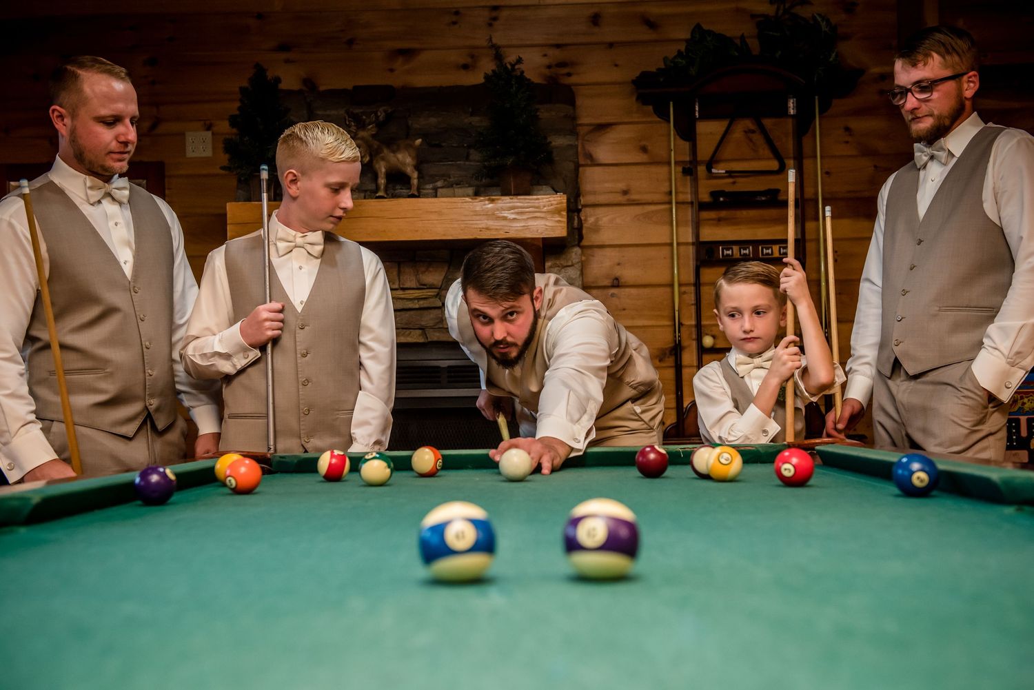 Groomsmen in tan vests playing pool on a green billiard table in a rustic wood-paneled room.