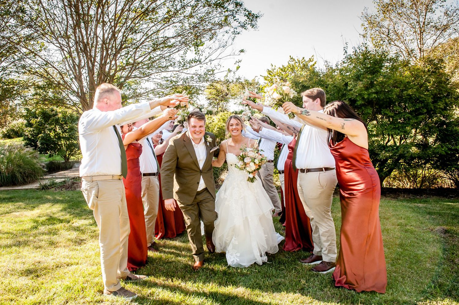 Wedding party in rustic attire celebrating couple walking under raised hands arch on sunny green lawn.