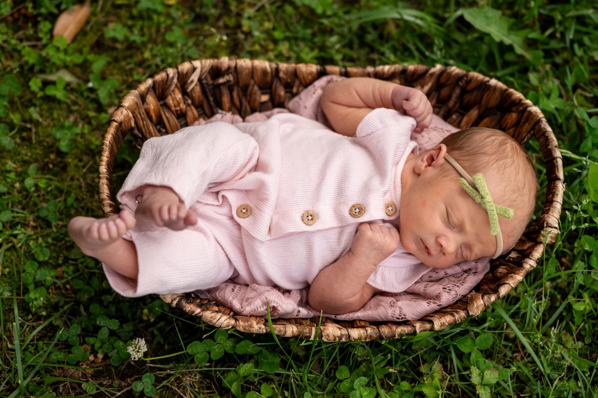 Sleeping baby in pink outfit lying in woven basket on grass.