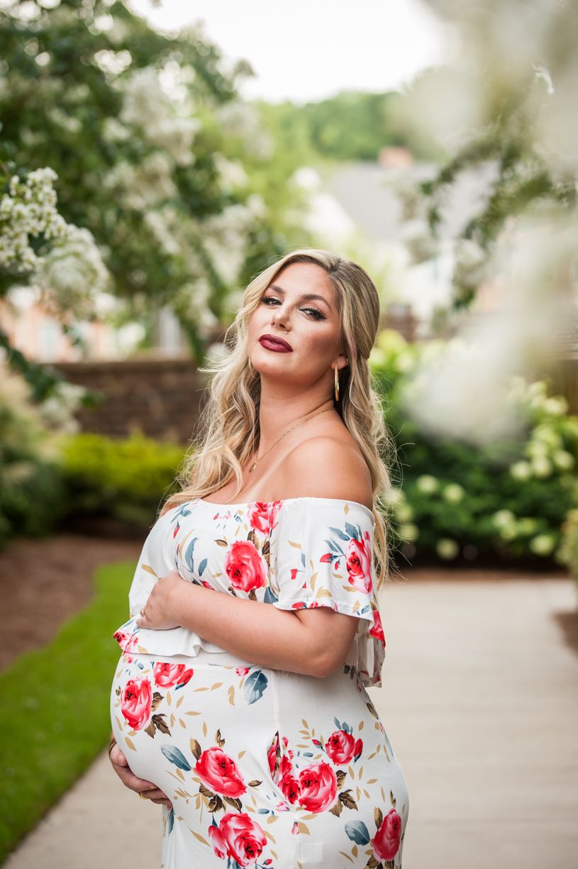 Maternity photo of person in floral dress posing in garden with blooming flowers.