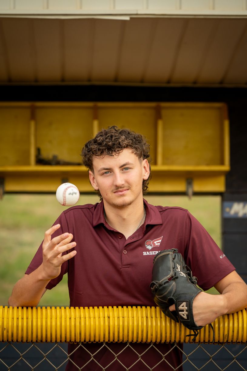 A senior baseball player poses with ball and glove in front of yellow dugout fence.