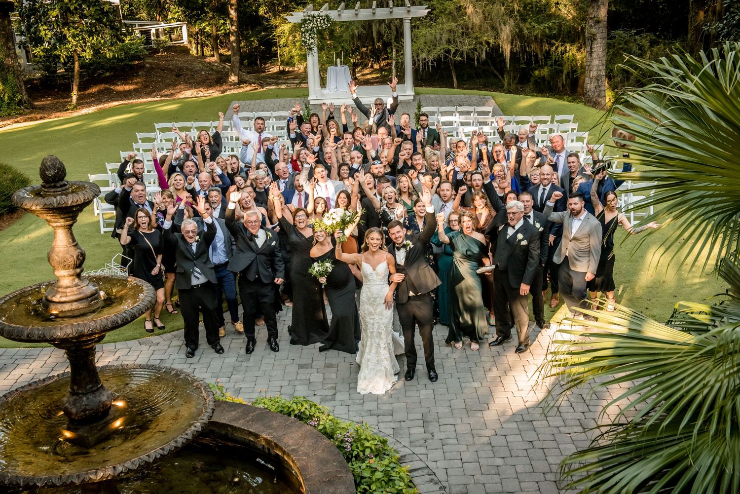 Aerial view of a large wedding group photo on a stone patio with fountain, surrounded by lush greenery.