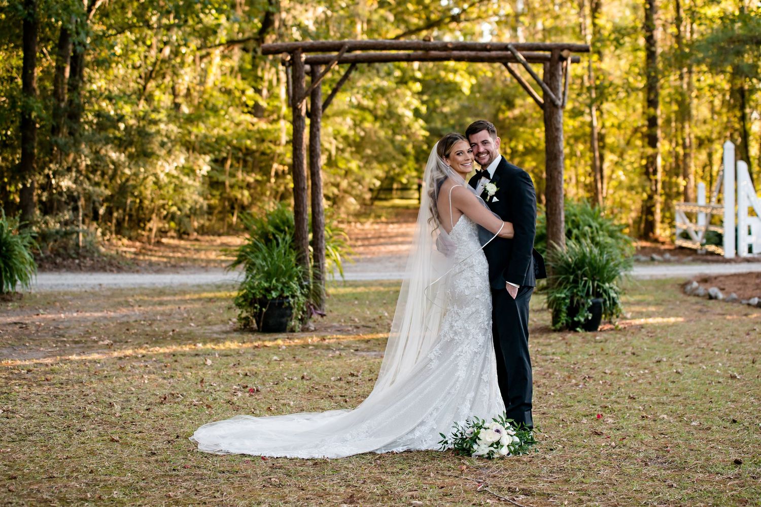 Bride and groom embrace outdoors under a wooden arbor surrounded by lush green trees in a romantic fall wedding setting.