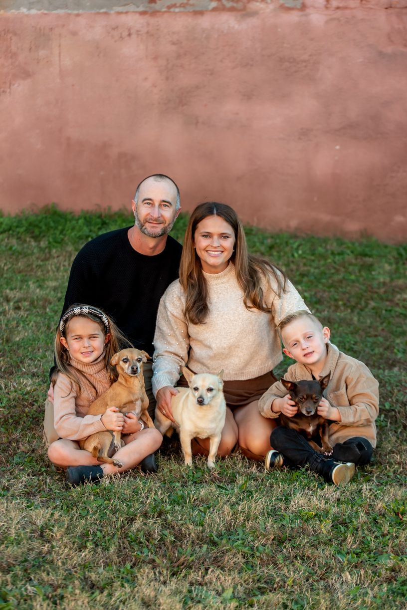A family sits together on grass with their three small dogs against a rustic pink wall.
