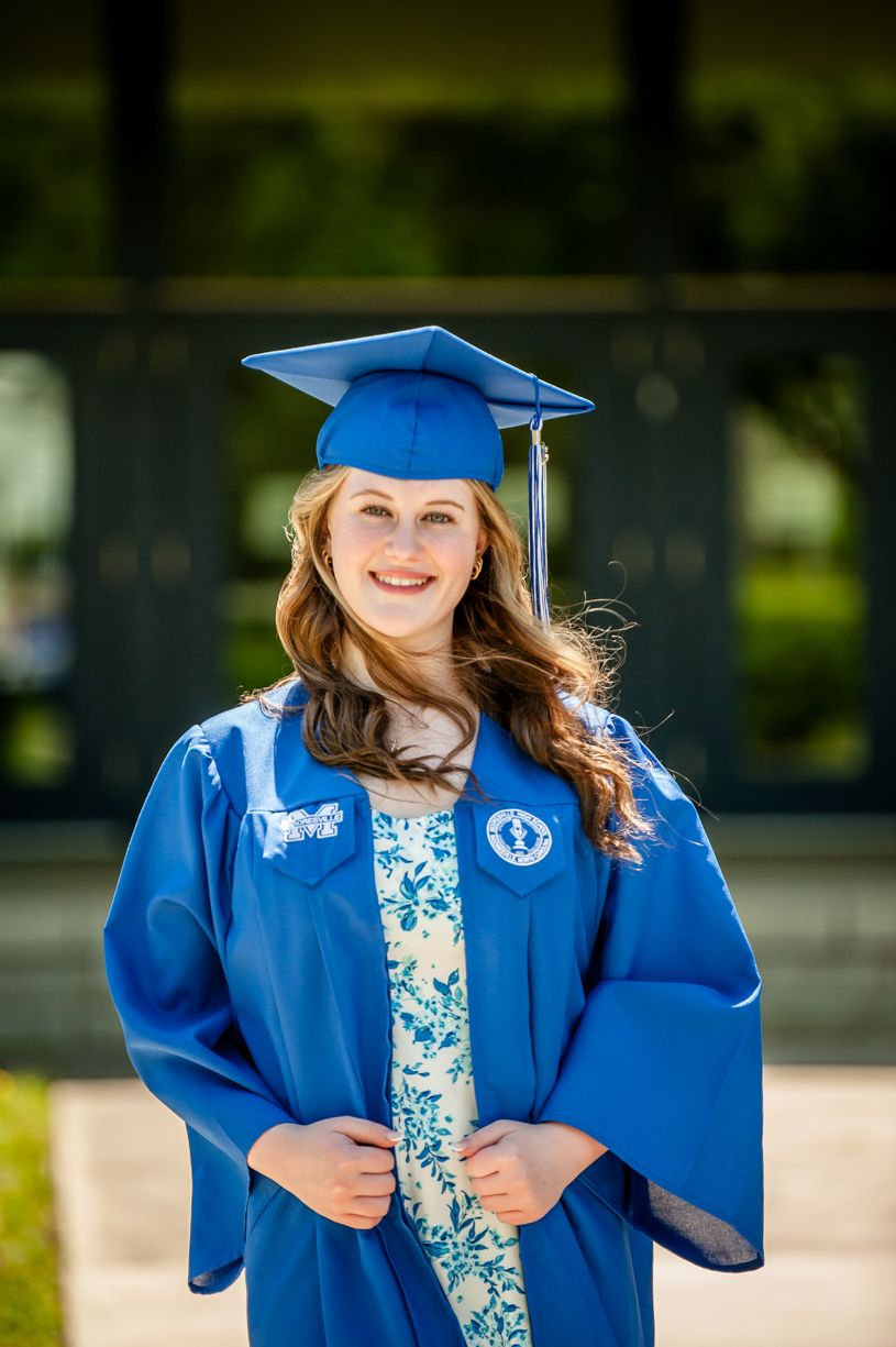 Graduate in blue cap and gown smiles during outdoor graduation ceremony.