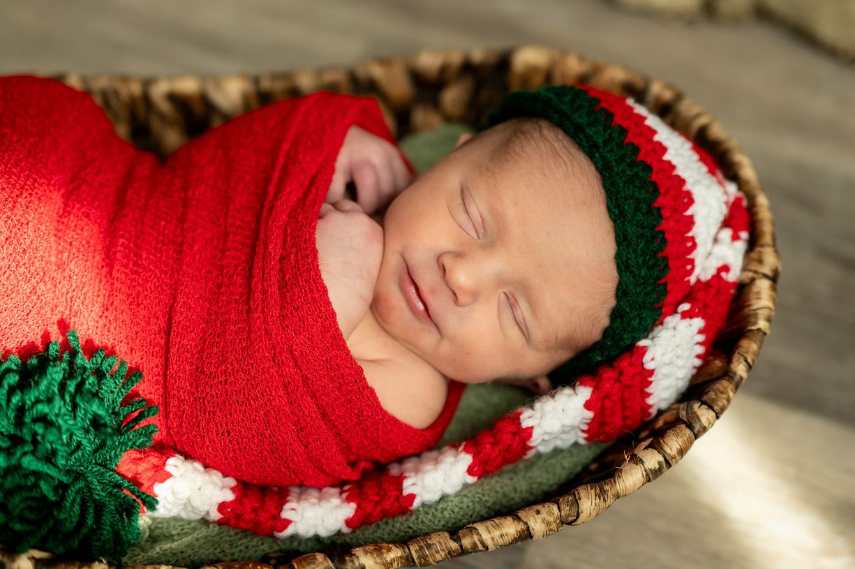 Sleeping newborn wrapped in red and white striped Christmas knit blanket with green pom detail in rustic basket.