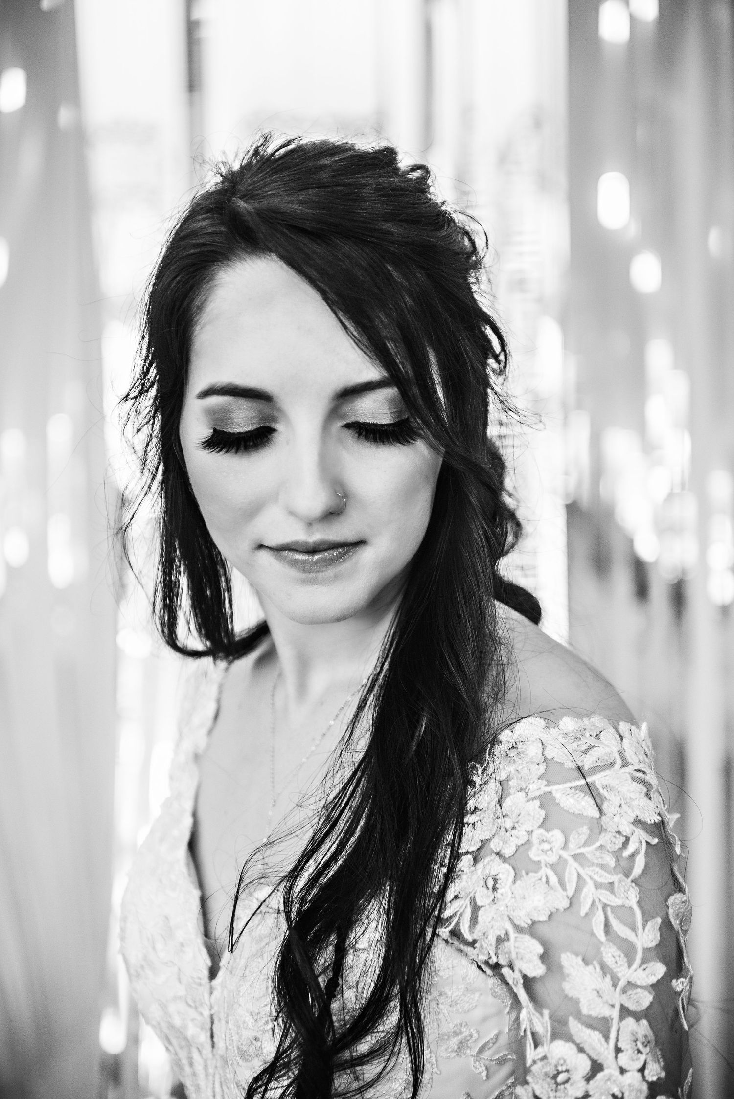 Black and white portrait of a bride with dark hair, lace off-shoulder gown, and elegant makeup, eyes downcast.